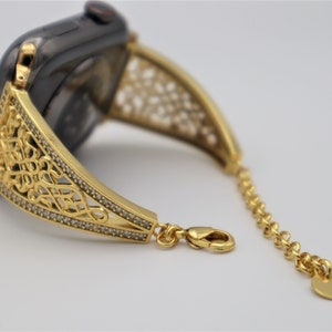 May include: A gold-toned watch band with a delicate, intricate design. The band is adorned with small, clear stones and features a chain link extension.
