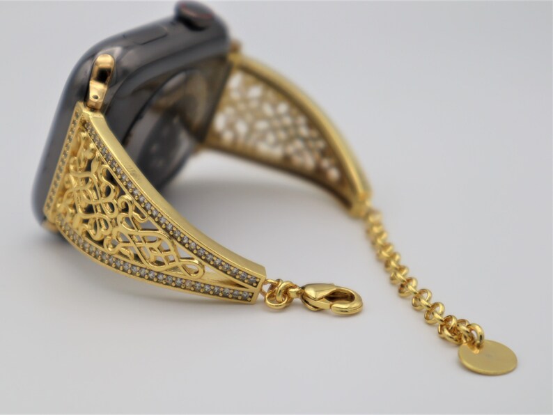 May include: A gold-toned watch band with a delicate, intricate design. The band is adorned with small, clear stones and features a chain link extension.
