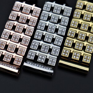May include: Close-up of three metal watch straps in rose gold, silver, and gold. Each strap features a repeating pattern of square links encrusted with small, clear gemstones. The straps are displayed against a dark background.