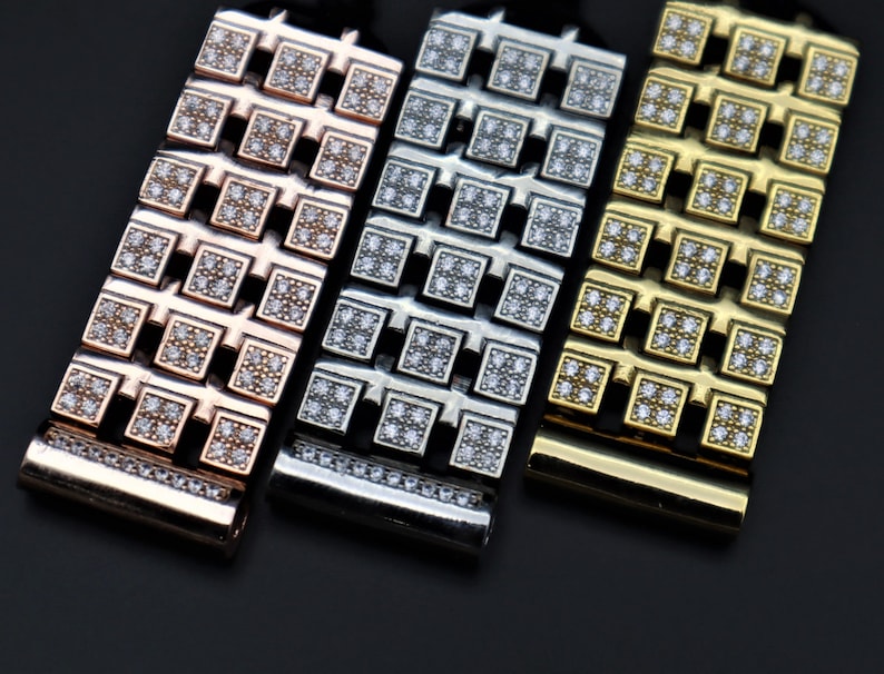 May include: Close-up of three metal watch straps in rose gold, silver, and gold. Each strap features a repeating pattern of square links encrusted with small, clear gemstones. The straps are displayed against a dark background.