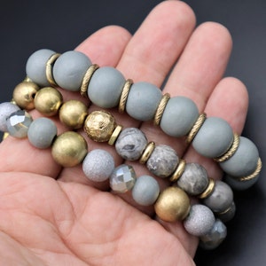 May include: A set of three bracelets featuring gray wooden beads, gold beads, and gray and white marbled beads.