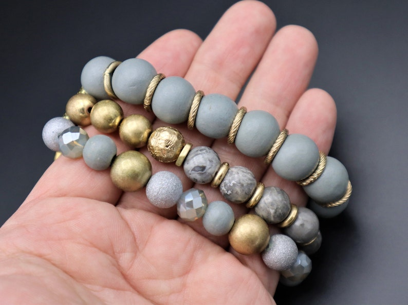 May include: A set of three bracelets featuring gray wooden beads, gold beads, and gray and white marbled beads.