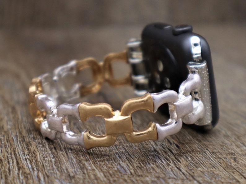 May include: A two-tone watch band with a chain link design. The links alternate between silver and gold-colored metal. The band is attached to a black smartwatch, showcasing a modern and stylish accessory.