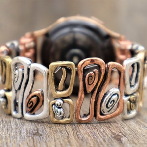 May include: A mixed-metal bracelet with a unique design. The bracelet features silver, gold, and copper-toned links with abstract shapes and spiral details. The bracelet is shown on a wooden surface.