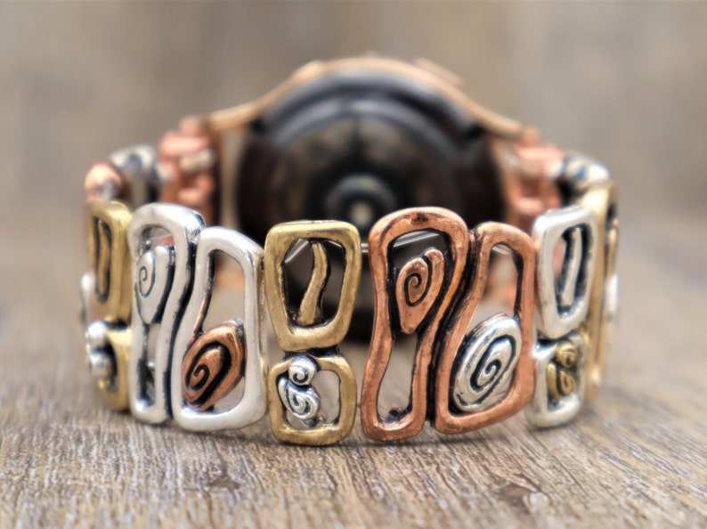 May include: A mixed-metal bracelet with a unique design. The bracelet features silver, gold, and copper-toned links with abstract shapes and spiral details. The bracelet is shown on a wooden surface.