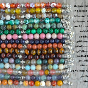 May include: A close-up of 15 strands of gemstone beads, each with a silver clasp. The beads are arranged in rows, with each row representing a different type of gemstone. The gemstones include fire agate, olive agate, coffee agate, green agate, African turquoise, malachite, rhodonite, blue-sand stone, golden-sand stone, yellow tiger's eye, red tiger's eye, Indian agate, amazonite, volcano cherry quartz, fluorite, and mookaite.