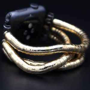 Puede incluir: Una pulsera de reloj de metal dorado envuelve un reloj inteligente negro. La pulsera está hecha de múltiples hebras de metal entrelazadas.