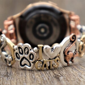 May include: A copper and silver watch band with charms including a paw print, a heart, the words "I love cats", and a cat silhouette.