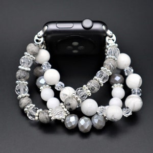 May include: A silver and white beaded Apple Watch band with grey, white, and clear beads. The band is made of multiple strands of beads and is designed to fit around the watch.
