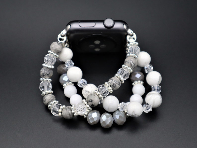 May include: A silver and white beaded Apple Watch band with grey, white, and clear beads. The band is made of multiple strands of beads and is designed to fit around the watch.