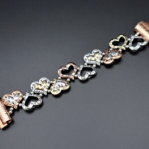 May include: A silver, gold, and copper bracelet with a series of heart-shaped charms. The bracelet has a clasp on each end.