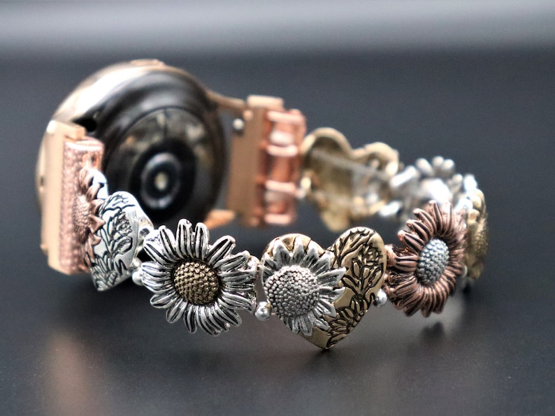 May include: A rose gold smartwatch band with a decorative sunflower and heart design. The band is made of metal and has a clasp closure.