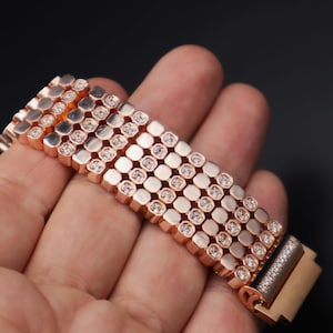 May include: A rose gold bracelet with a row of square-shaped stones set in a repeating pattern. The bracelet is made of metal and has a polished finish.