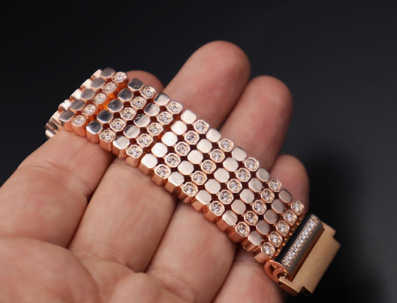 May include: A rose gold bracelet with a row of square-shaped stones set in a repeating pattern. The bracelet is made of metal and has a polished finish.