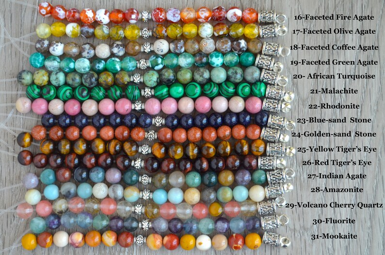 May include: A close-up of 15 different gemstone bracelets with silver clasps. Each bracelet is labeled with the type of gemstone, including Fire Agate, Olive Agate, Coffee Agate, Green Agate, African Turquoise, Malachite, Rhodonite, Blue-sand Stone, Golden-sand Stone, Yellow Tiger's Eye, Red Tiger's Eye, Indian Agate, Amazonite, Volcano Cherry Quartz, Fluorite, and Mookaite.