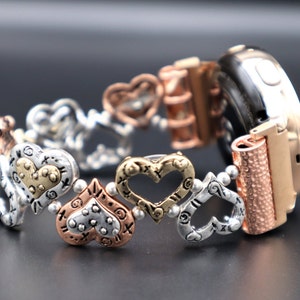 May include: A rose gold smartwatch band with a silver and gold heart charm bracelet. The bracelet features a variety of heart designs, including open hearts, hearts with dots, and hearts with a textured surface.