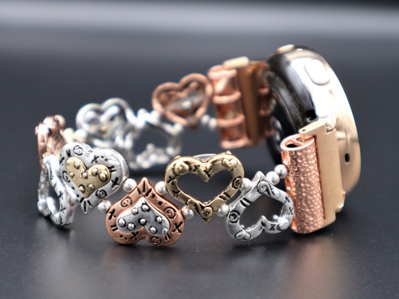 May include: A rose gold smartwatch band with a silver and gold heart charm bracelet. The bracelet features a variety of heart designs, including open hearts, hearts with dots, and hearts with a textured surface.