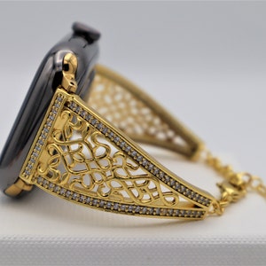May include: A gold-toned watch band with a delicate, intricate design and clear crystal accents. The band is attached to a black smartwatch.