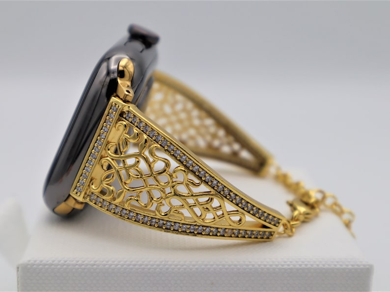 May include: A gold-toned watch band with a delicate, intricate design and clear crystal accents. The band is attached to a black smartwatch.