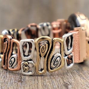 May include: A multi-toned bracelet with a mix of copper, silver, and gold-colored metal links. The links feature abstract swirl and geometric designs. The watch face is a rose gold color with a textured finish.