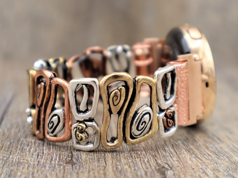 May include: A multi-toned bracelet with a mix of copper, silver, and gold-colored metal links. The links feature abstract swirl and geometric designs. The watch face is a rose gold color with a textured finish.