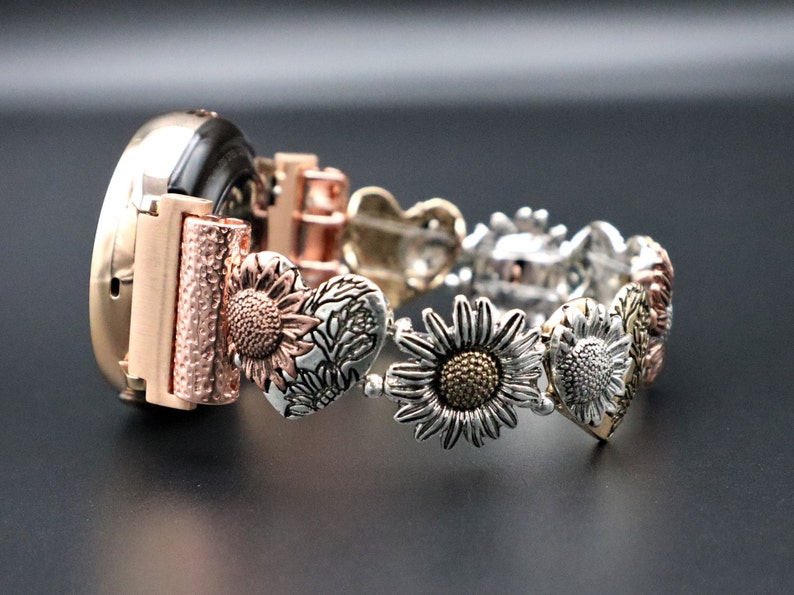 May include: A rose gold watch band with a silver and gold sunflower design. The band is made of metal and has a heart-shaped design.