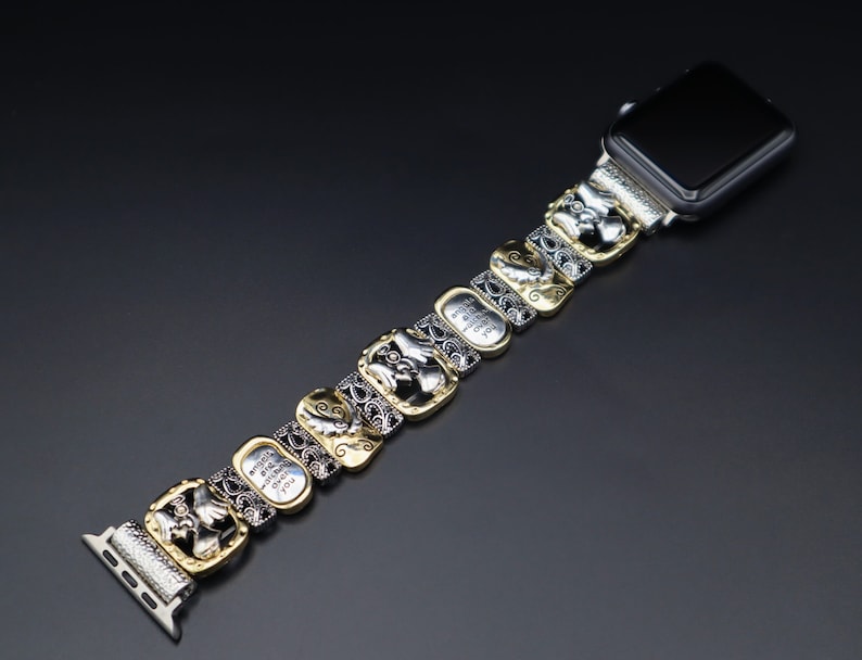 May include: A silver and gold watch band with a decorative design. The band is made up of multiple links, each with a different design. The band is attached to a black Apple Watch.