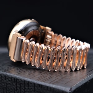 K&ouml;nnte beinhalten: Eine goldfarbene Armbanduhr mit einem runden Zifferblatt und einem einzigartigen, filigranen Metallband. Die Uhr liegt auf einer schwarzen Oberfl&auml;che.