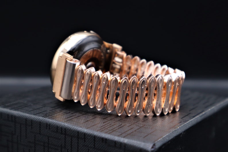 K&ouml;nnte beinhalten: Eine goldfarbene Armbanduhr mit einem runden Zifferblatt und einem einzigartigen, filigranen Metallband. Die Uhr liegt auf einer schwarzen Oberfl&auml;che.