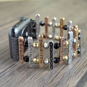 Puede incluir: Correa de Apple Watch multicolor con barras metálicas verticales grabadas con las palabras "Incredible", "Spectacular", "Marvelous", "Phenomenal" y "Fantastic". La correa presenta detalles metálicos en negro, dorado, plateado y cobrizo, y cuentas negras.