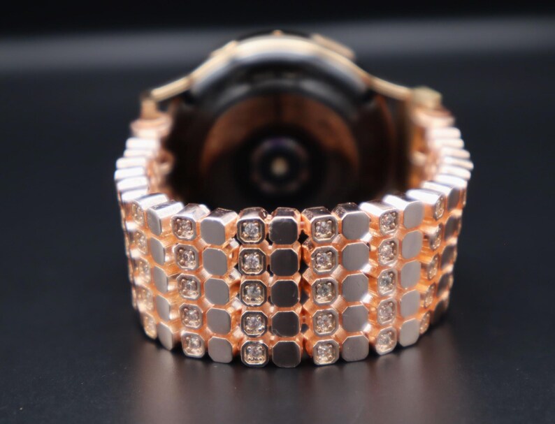 May include: A rose gold smartwatch band with a diamond-like pattern. The band is made of metal and has a polished finish.
