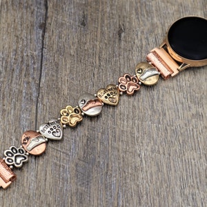 Puede incluir: Un reloj inteligente de oro rosa con una correa decorativa con huellas de patas y corazones. La correa tiene una mezcla de dijes dorados, plateados y cobrizos, algunos con la inscripci&oacute;n "Best Friend".