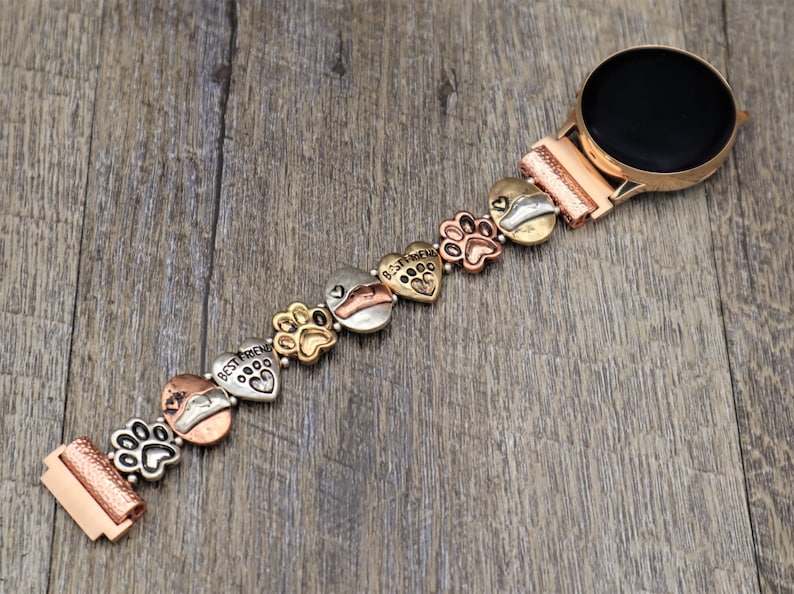 Puede incluir: Un reloj inteligente de oro rosa con una correa decorativa con huellas de patas y corazones. La correa tiene una mezcla de dijes dorados, plateados y cobrizos, algunos con la inscripci&oacute;n "Best Friend".
