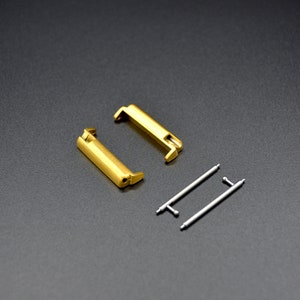 May include: Two gold-colored watch band connectors and two silver spring bars. The connectors are rectangular with rounded edges. The spring bars are straight and have a small loop at each end.
