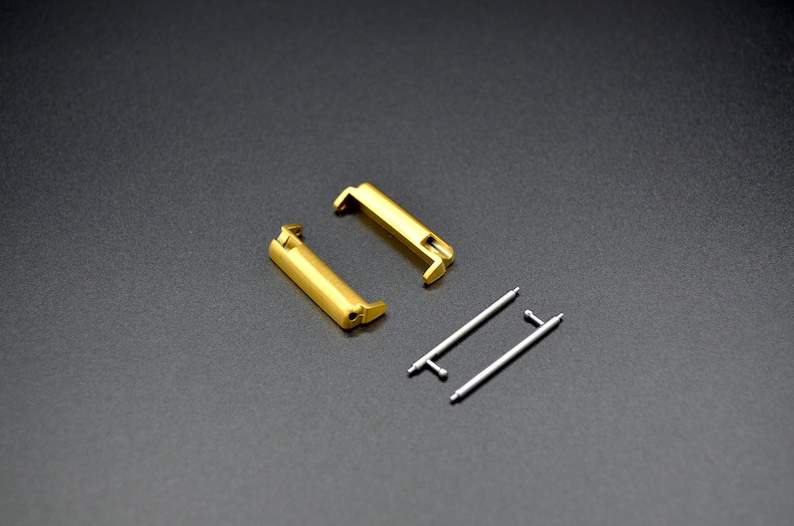 May include: Two gold-colored watch band connectors and two silver spring bars. The connectors are rectangular with rounded edges. The spring bars are straight and have a small loop at each end.