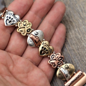 Puede incluir: Pulsera de metal multicolor con dijes en forma de coraz&oacute;n con el texto "BEST FRIEND" y huellas de patas. Otros dijes incluyen dise&ntilde;os de huesos de perro y caras de perro.
