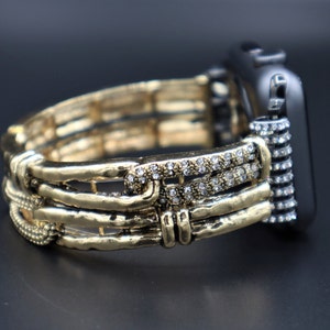 May include: A gold and silver watch band with a crystal embellishment. The band is made of multiple layers of metal and has a textured finish.