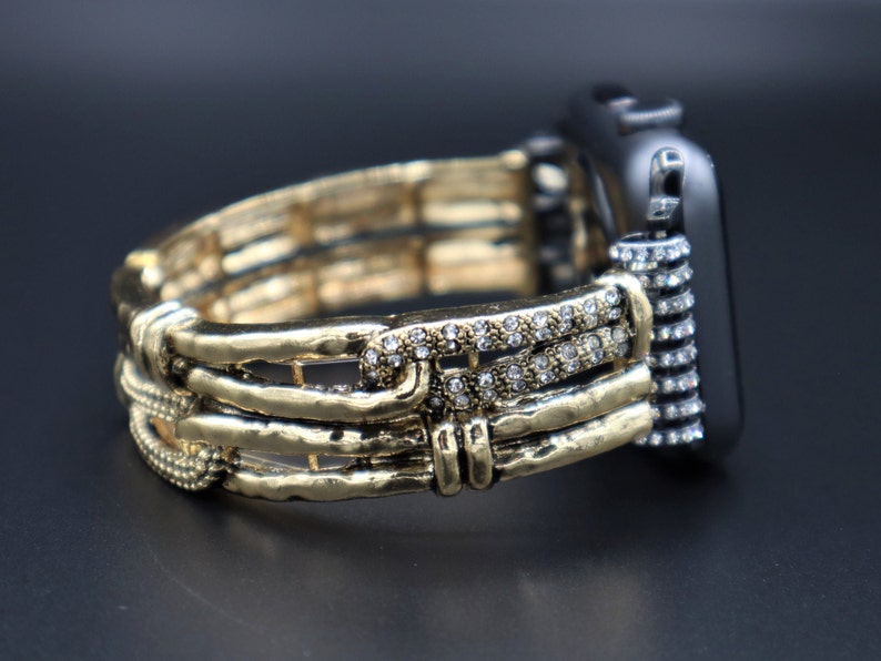 May include: A gold and silver watch band with a crystal embellishment. The band is made of multiple layers of metal and has a textured finish.