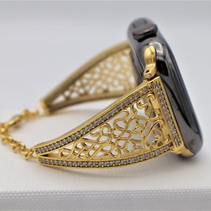 May include: A gold-tone watch band with a decorative, intricate design and clear crystals. The band is attached to a black smartwatch with a gold-tone digital display.