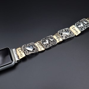 May include: A silver and gold Apple Watch band with a heart charm and the words "Love", "Faith", "Hope", and "Love" engraved on each charm.