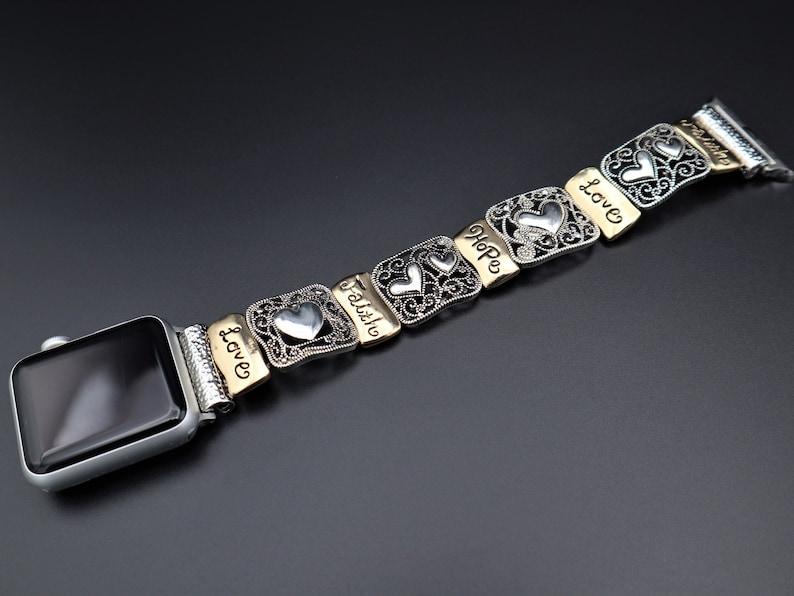 May include: A silver and gold Apple Watch band with a heart charm and the words "Love", "Faith", "Hope", and "Love" engraved on each charm.