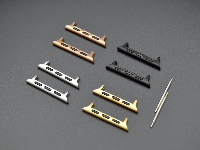May include: An assortment of watch band adapters in various metallic finishes, including gold, silver, bronze, and black. The adapters have a rectangular shape with a series of small, rectangular cutouts and are accompanied by two small silver pins.
