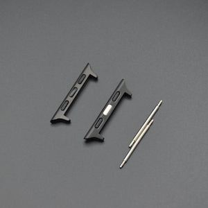 May include: Two black watch band adapters and two silver spring bars are displayed on a dark gray surface. The adapters have a rectangular shape with three small holes and a curved end. The spring bars are thin, cylindrical, and have a pin on each end.