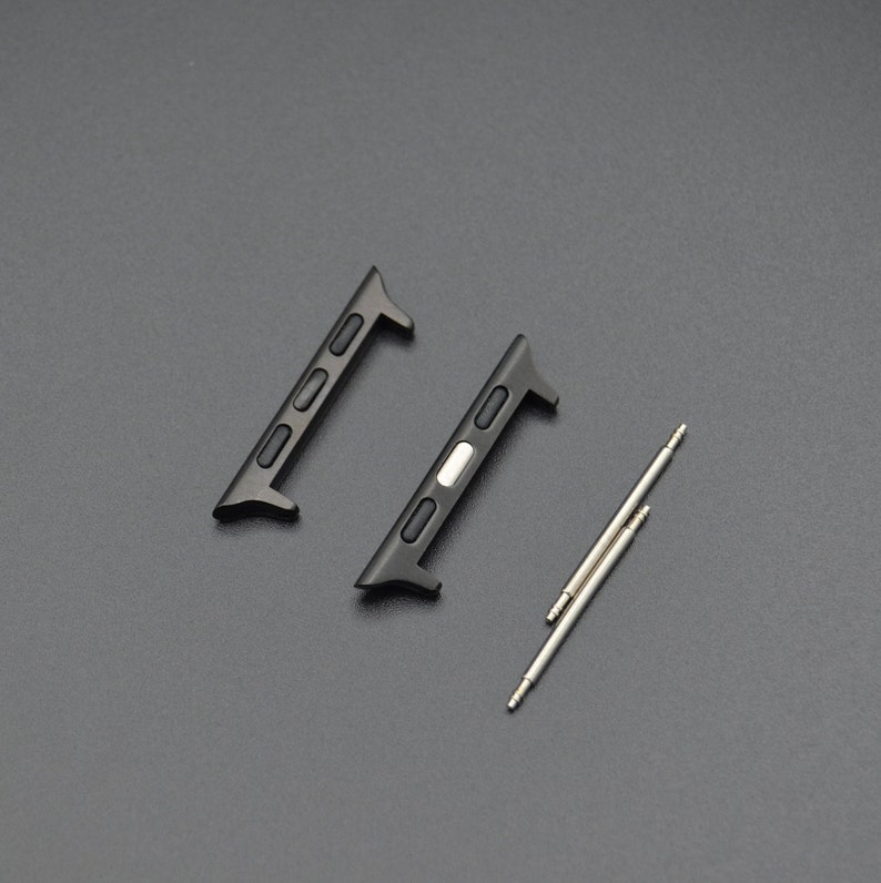 May include: Two black watch band adapters and two silver spring bars are displayed on a dark gray surface. The adapters have a rectangular shape with three small holes and a curved end. The spring bars are thin, cylindrical, and have a pin on each end.