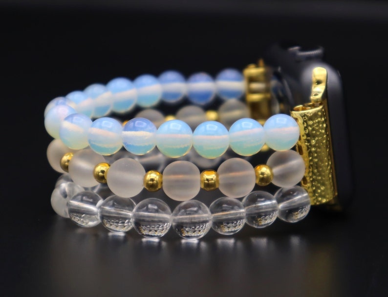 K&ouml;nnte beinhalten: Ein goldenes Apple Watch Armband mit drei Perlenreihen. Die oberste Reihe besteht aus hellblauen Opalperlen, die mittlere Reihe aus matten wei&szlig;en Perlen und die unterste Reihe aus klaren Glasperlen.