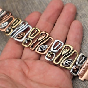 May include: A multi-tone metal bracelet with copper, silver, and gold-colored links. The bracelet features an openwork design with geometric shapes and spiral details. The clasp is a matching copper color.