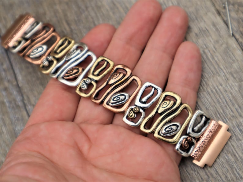 May include: A multi-tone metal bracelet with copper, silver, and gold-colored links. The bracelet features an openwork design with geometric shapes and spiral details. The clasp is a matching copper color.