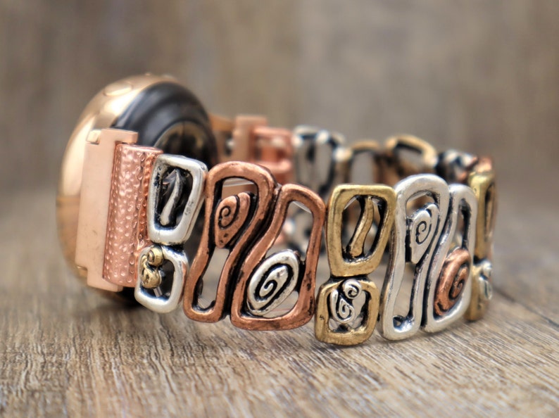 May include: A multi-toned metal bracelet with a watch face. The bracelet features a mix of copper, silver, and gold-colored links with an open, geometric design. The watch face is rose gold.
