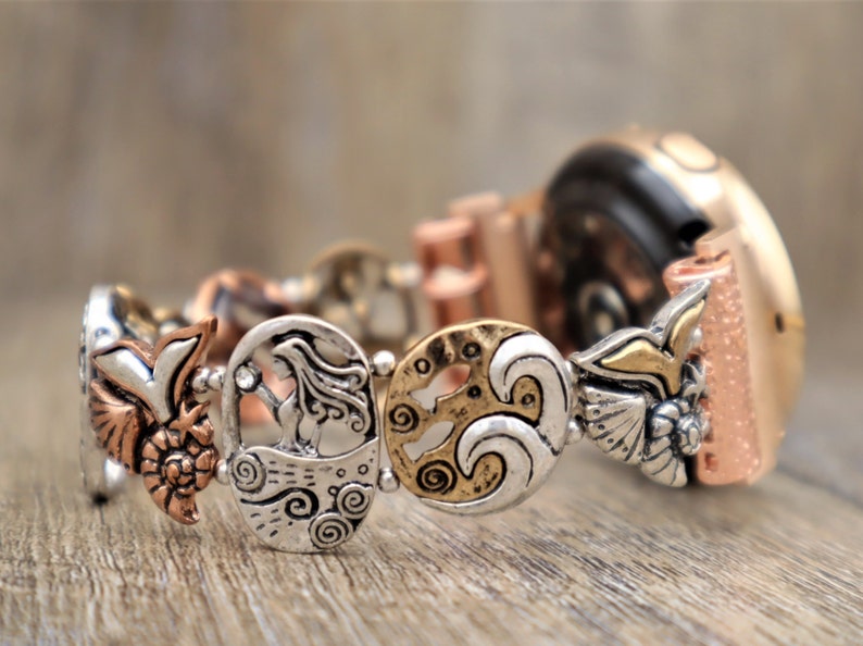 May include: A silver and gold watch band with a mermaid, wave, and bird design. The band is made of metal and has a textured finish.
