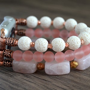Puede incluir: Una pulsera de Apple Watch de tres hebras hecha con cuentas de roca de lava blanca, cuentas de vidrio rosa y cuentas de cuarzo rosa. La pulsera presenta detalles en cobre.